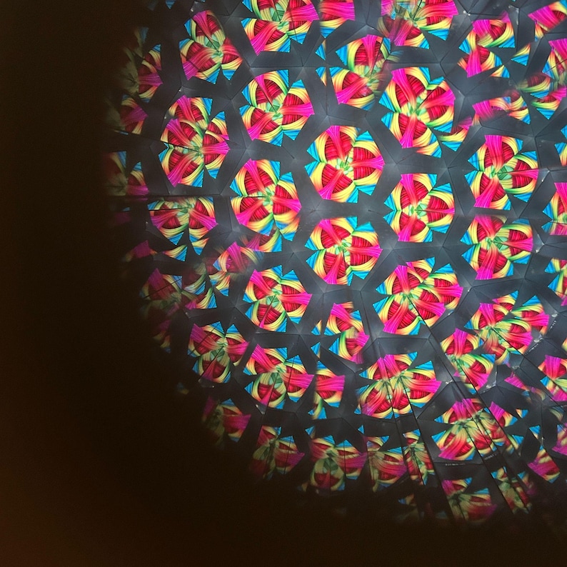 May include: Kaleidoscope image with a repeating pattern of colorful, geometric shapes. The design features vibrant pink, yellow, and blue hues against a dark background. The pattern creates a symmetrical, abstract design, reminiscent of a flower or star.