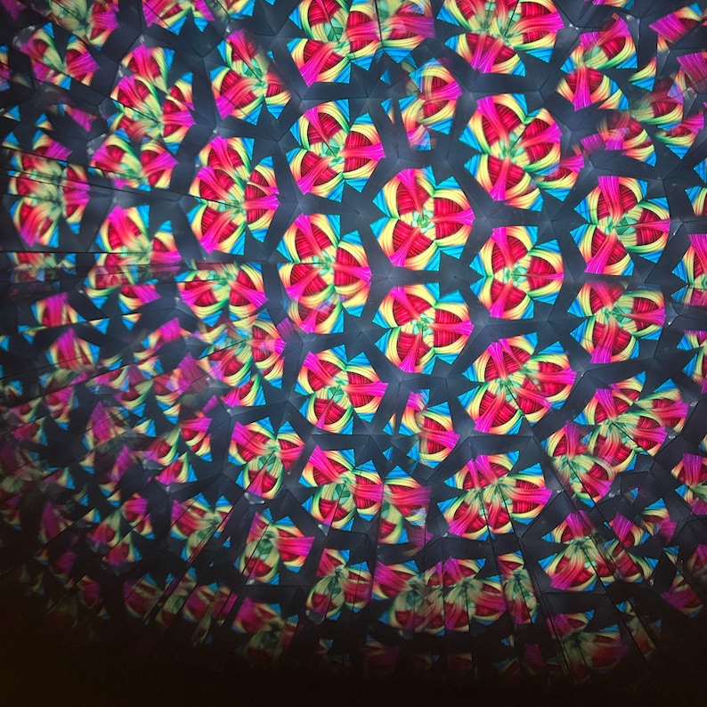 May include: A kaleidoscope view of a colorful pattern. The image features a repeating design of geometric shapes in shades of pink, yellow, blue, and black. The pattern creates a symmetrical, abstract design.