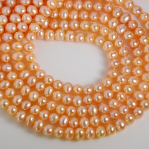 May include: A strand of peach-coloured freshwater pearls. The pearls are round and smooth, and they are strung on a white thread. The pearls are arranged in a loose, flowing pattern.