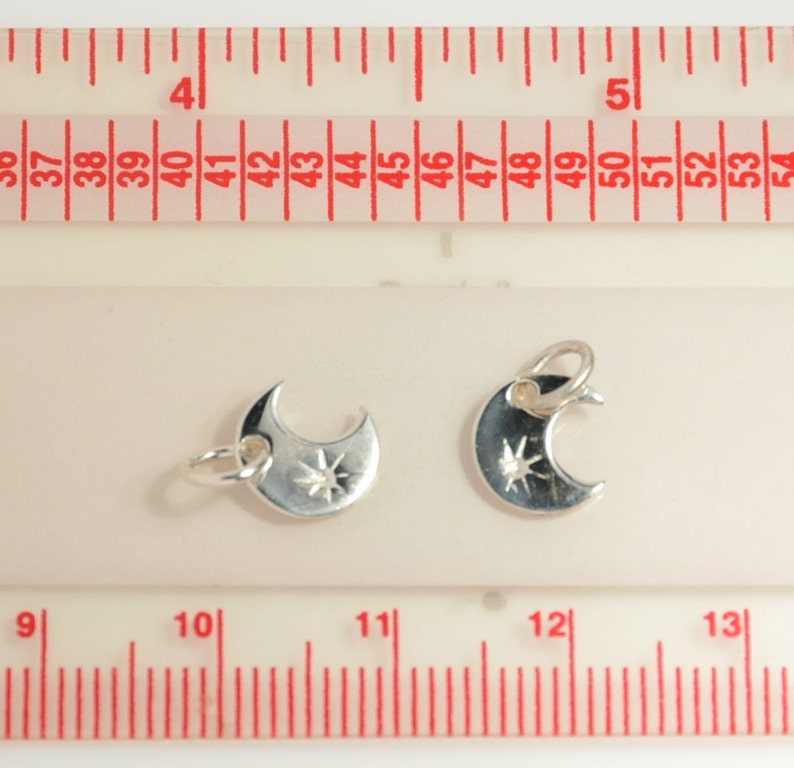 May include: Two silver crescent moon charms with a star design. The charms are approximately 3.2 cm in length and are shown against a white background with a ruler for scale.