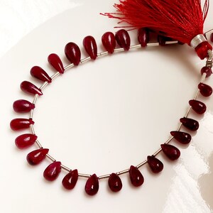May include: A strand of deep red ruby gemstone beads in a teardrop shape. The beads are strung on silver wire and have a tassel at the end.