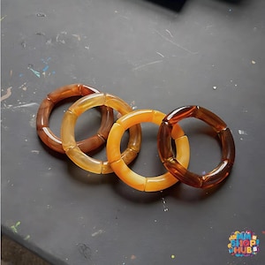 Op de afbeelding: Vier doorschijnende, dikke armbanden in bruin- en amberkleuren. De armbanden zijn gerangschikt op een donkere ondergrond, waarbij hun verschillende tinten en geometrische ontwerp worden getoond. Elke armband bestaat uit rechthoekige segmenten.