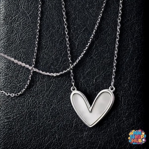 May include: A silver necklace with a heart-shaped pendant. The pendant has a white, pearlescent center and a silver border. The necklace chain is delicate and hangs on a black background.