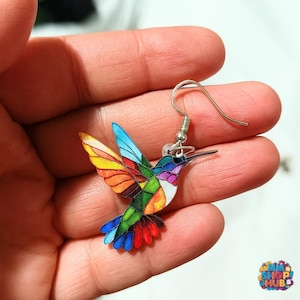 Puede incluir: Un pendiente de colibrí colorido con un gancho plateado. El pájaro presenta una vibrante gama de colores, incluyendo azul, naranja, verde y rojo. El pendiente es una representación artística detallada de un colibrí.