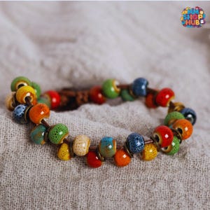 May include: A colorful ceramic bead bracelet. The bracelet features a variety of round beads in shades of red, orange, yellow, green, and blue. The beads are strung on a brown cord and are accented with gold-colored metal rings.