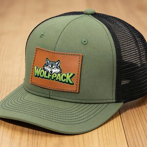 May include: An olive green baseball cap with a black mesh back and a flat brim. A rectangular brown leather patch is stitched to the front, featuring a graphic of a wolf head and the text "WOLF PACK" in bold green letters with a black outline.