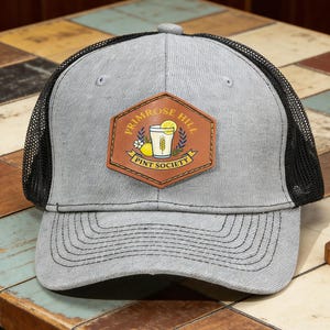 May include: A gray trucker hat with a black mesh back and a brown leather patch. The patch features the words "Primrose Hill Pint Society" around a graphic of a drink with a lemon. A small wooden deer figurine sits nearby.