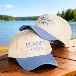 May include: Two baseball caps with a cream-colored crown and a light blue brim are stacked on a wooden surface. The caps have the embroidered text "SUMMER VIBES" in light blue. The background features a lake and trees.