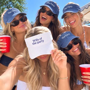 May include: A collection of five sun visors, four in blue denim with the white embroidered text The Party and one in white with the blue embroidered text Wife of the party. Each visor features a curved brim and an adjustable strap design.