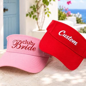 May include: A light pink visor with the embroidered text club Bride in a dark pink script font and a bright red visor with the embroidered text Custom in a white script font. Both visors feature a curved brim and an adjustable back strap.