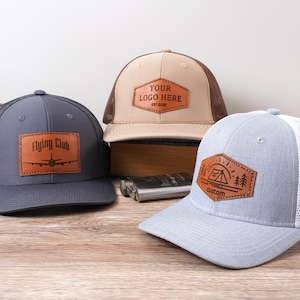 May include: Three trucker caps with leather patches. One is grey with a "Flying Club" patch, another is beige with a "YOUR LOGO HERE" patch, and the third is light blue with a camping scene patch. All have mesh backs.