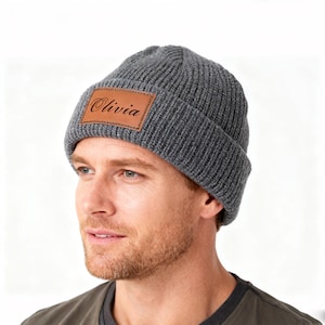 Peut inclure: Un bonnet en tricot gris avec un patch en cuir marron. Le patch est rectangulaire et porte le nom "Olivia" en cursive. Le bonnet est fait d'un matériau en tricot côtelé.