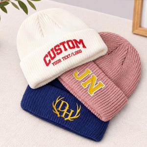 May include: Three ribbed knit beanies stacked together. The top white beanie features red embroidered text reading CUSTOM YOUR TEXT/LOGO. The middle pink beanie has yellow embroidered letters JN. The bottom blue beanie has a yellow embroidered DH logo.