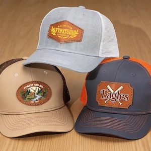 May include: Three trucker hats with leather patches. One is light gray with a white mesh back and a patch that says "VYNNSTUDIO". Another is tan with a brown mesh back and a "FOREST GUARD" patch. The third is navy blue with an orange mesh back and an "Eagles" patch.