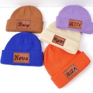 Peut inclure: Cinq bonnets en tricot de couleurs variées : marron, bleu, violet, crème et orange. Chaque bonnet est orné d'un patch rectangulaire en cuir avec un nom : "Daisy", "Nova", "Betty", "Ryan" et "Eliza". Les bonnets sont posés sur une surface blanche.