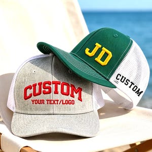 Op de afbeelding: Een grijs gemêleerde baseballpet met witte mesh achterpanelen en de rode geborduurde tekst CUSTOM YOUR TEXT/LOGO, gecombineerd met een groene baseballpet met gele geborduurde JD-letters en het zwarte woord CUSTOM aan de zijkant.