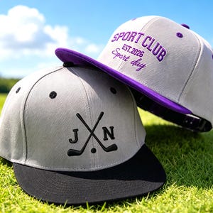 Op de afbeelding: Twee grijze baseballpetten met een platte klep. De voorste pet heeft zwarte geborduurde gekruiste golfclubs met de letters J en N. De achterste pet heeft paars borduurwerk met de tekst SPORT CLUB EST.2026 Sport day en een paarse onderkant.
