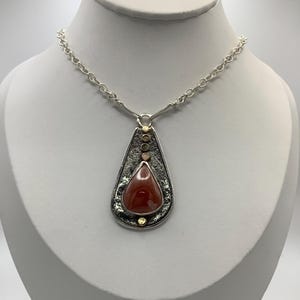 May include: A teardrop-shaped pendant necklace with a red agate stone set in a silver frame. The frame has a textured finish and is accented with small gold beads.