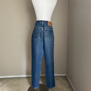Peut inclure: Un jean bleu denim avec un patch en cuir marron au dos. Le jean est sur un mannequin et a une coupe droite.