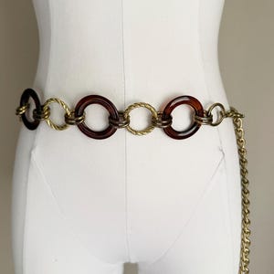May include: A vintage belt featuring a series of large, dark brown circular links alternating with smaller, gold-toned rope-like links. A gold chain dangles from the end. The belt is displayed on a white mannequin.