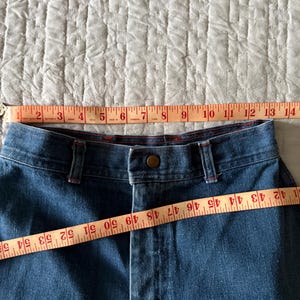 K&ouml;nnte beinhalten: Nahaufnahme einer blauen Jeans mit Knopfverschluss und G&uuml;rtelschlaufen. Ein Ma&szlig;band zeigt, dass die Bundweite etwa 33 cm betr&auml;gt. Die Jeans hat rote Nahtdetails.
