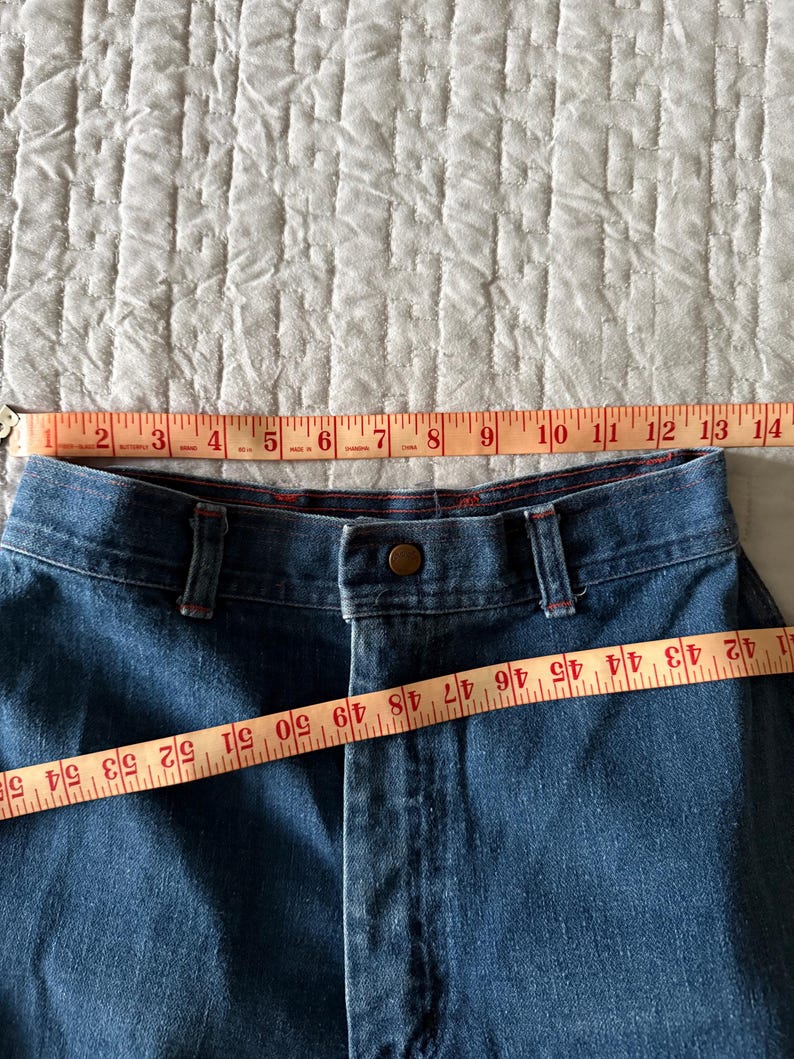 K&ouml;nnte beinhalten: Nahaufnahme einer blauen Jeans mit Knopfverschluss und G&uuml;rtelschlaufen. Ein Ma&szlig;band zeigt, dass die Bundweite etwa 33 cm betr&auml;gt. Die Jeans hat rote Nahtdetails.