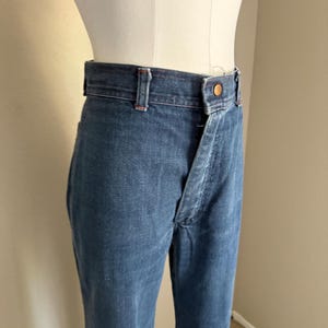 K&ouml;nnte beinhalten: Blaue Jeans aus Denim mit hoher Taille und Knopfverschluss. Die Jeans haben ein klassisches F&uuml;nf-Taschen-Design und ein gerades Bein. Der Denim hat ein leicht verwaschenes Aussehen.
