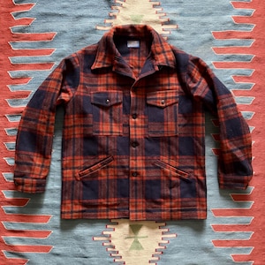 May include: A vintage-style, button-down flannel shirt in a red and navy plaid pattern. The shirt has two chest pockets and two lower pockets. The collar is a classic spread collar. The shirt is laid flat on a patterned rug.