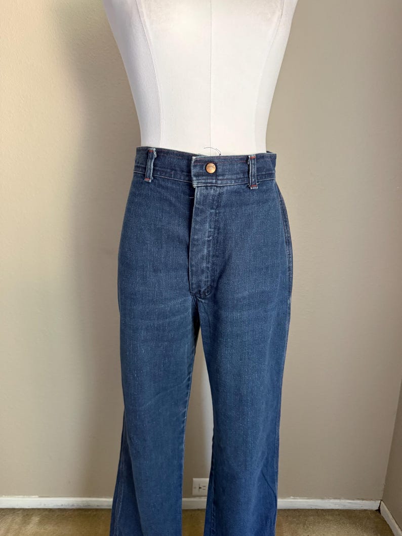 K&ouml;nnte beinhalten: Dunkelblaue Jeans mit hoher Taille und ausgestellten Beinen. Die Jeans haben einen Knopfverschluss und ein klassisches F&uuml;nf-Taschen-Design. Die Jeans werden auf einer Schaufensterpuppe pr&auml;sentiert.