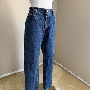 May include: High-waisted dark blue denim jeans featuring a classic five-pocket design, a metal button closure, and a zip fly. The jeans have a straight-leg cut with visible stitching along the seams and a standard waistband with belt loops.