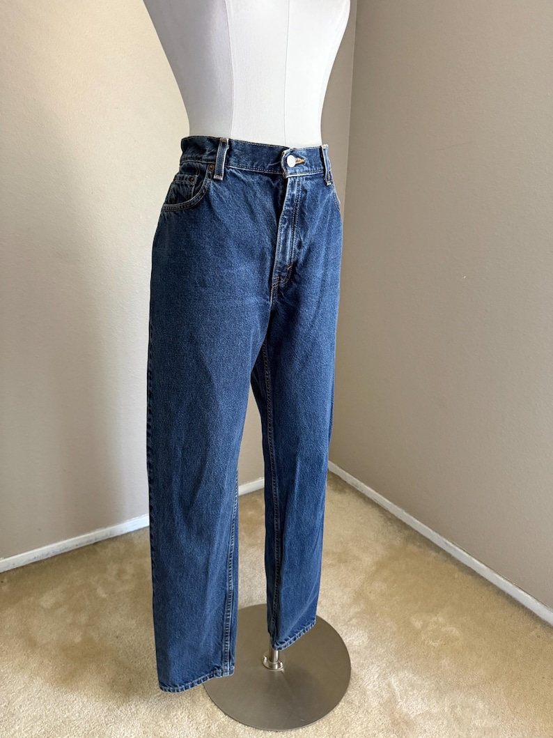 May include: High-waisted dark blue denim jeans featuring a classic five-pocket design, a metal button closure, and a zip fly. The jeans have a straight-leg cut with visible stitching along the seams and a standard waistband with belt loops.