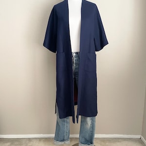 May include: A navy blue, open-front kimono-style jacket with short sleeves and two front pockets. The jacket is long, reaching to the mid-calf, and is displayed on a mannequin with blue jeans. The garment has side slits.