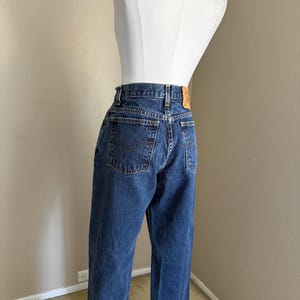 May include: Dark blue denim jeans featuring a high-waisted fit, a classic five-pocket design with stitched arcuate detailing on the back pockets, a tan leather-like patch on the waistband, and a small red tab on the right rear pocket seam.