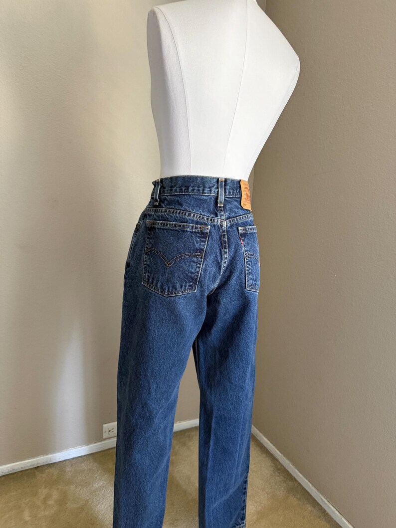 May include: Dark blue denim jeans featuring a high-waisted fit, a classic five-pocket design with stitched arcuate detailing on the back pockets, a tan leather-like patch on the waistband, and a small red tab on the right rear pocket seam.