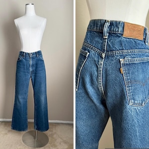 May include: High-waisted blue denim jeans featuring a straight-leg cut, a tan leather patch on the back waistband, and an orange tab on the back pocket. The jeans have a classic five-pocket design with visible stitching and a button fly closure.