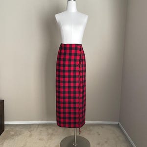 May include: A long wrap skirt featuring a red and black buffalo plaid pattern. The skirt has a straight silhouette, a high waist, and a vertical edge detail with fringe trim running down the front side. The garment is displayed on a mannequin.