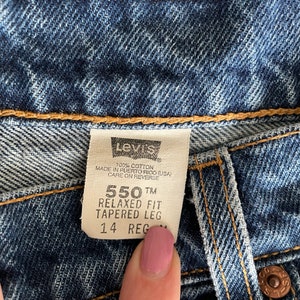 Peut inclure: Une &eacute;tiquette de jean Levi's 550 Relaxed Fit Tapered Leg en denim bleu avec du texte blanc. L'&eacute;tiquette indique "100% coton", "Fabriqu&eacute; &agrave; Porto Rico (USA)", "Entretien au dos", "550 TM", "Coupe d&eacute;contract&eacute;e", "Pantalon fusel&eacute;" et "14 Reg".