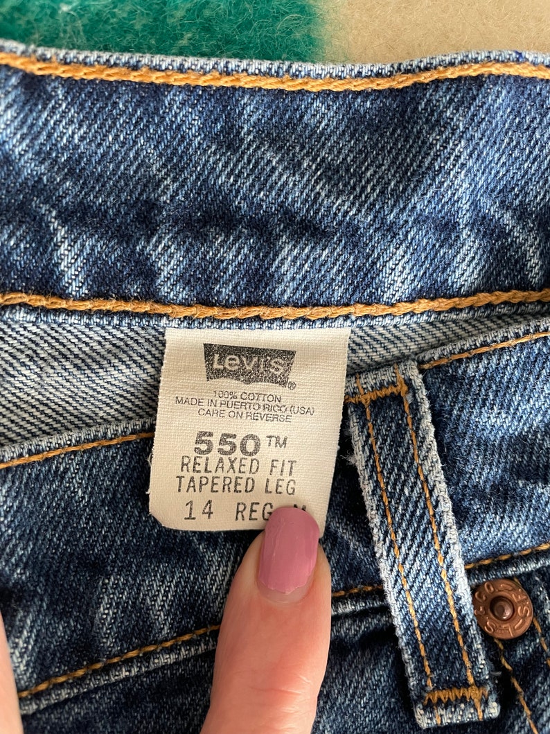 Peut inclure: Une &eacute;tiquette de jean Levi's 550 Relaxed Fit Tapered Leg en denim bleu avec du texte blanc. L'&eacute;tiquette indique "100% coton", "Fabriqu&eacute; &agrave; Porto Rico (USA)", "Entretien au dos", "550 TM", "Coupe d&eacute;contract&eacute;e", "Pantalon fusel&eacute;" et "14 Reg".