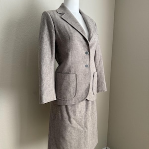 May include: A two-piece women's suit in a neutral taupe color. The suit includes a blazer with two front pockets and a matching knee-length skirt. The blazer has two dark buttons and a notched lapel. The suit is displayed on a mannequin.