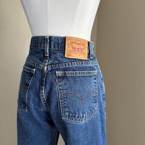 May include: Dark blue denim jeans featuring a high-waisted fit, belt loops, and two back pockets. A tan leather patch is stitched to the waistband with the text Levi Strauss & Co. Original Riveted Quality Clothing Every Garment Guaranteed 550.