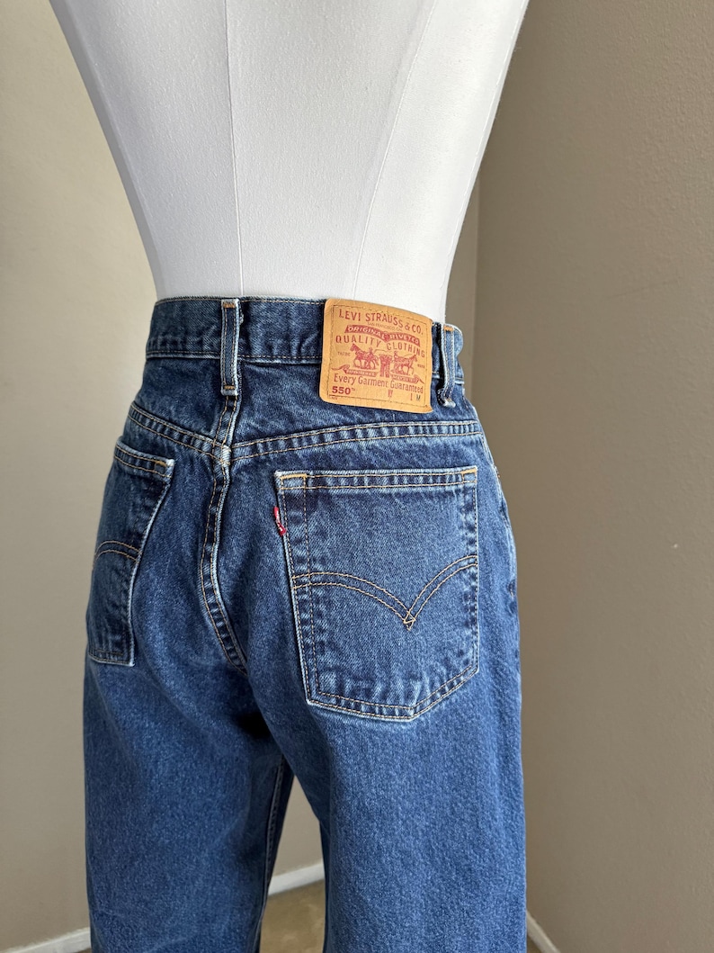May include: Dark blue denim jeans featuring a high-waisted fit, belt loops, and two back pockets. A tan leather patch is stitched to the waistband with the text Levi Strauss & Co. Original Riveted Quality Clothing Every Garment Guaranteed 550.