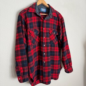 Puede incluir: Una camisa de franela a cuadros rojos y negros con cuello acolchado y dos bolsillos en el pecho. La camisa de manga larga es abotonada y cuelga de una percha de madera contra una pared blanca.