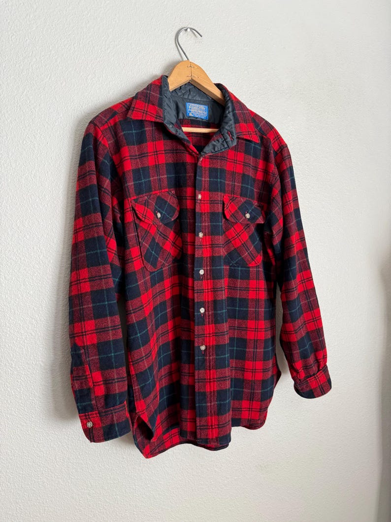 Puede incluir: Una camisa de franela a cuadros rojos y negros con cuello acolchado y dos bolsillos en el pecho. La camisa de manga larga es abotonada y cuelga de una percha de madera contra una pared blanca.