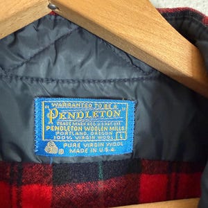 Puede incluir: Primer plano de una chaqueta de lana a cuadros rojos y negros. La chaqueta tiene un forro acolchado azul y una etiqueta azul que dice "WARRANTED TO BE A PENDLETON" y "PURE VIRGIN WOOL MADE IN U.S.A.". Cuelga de una percha de madera.