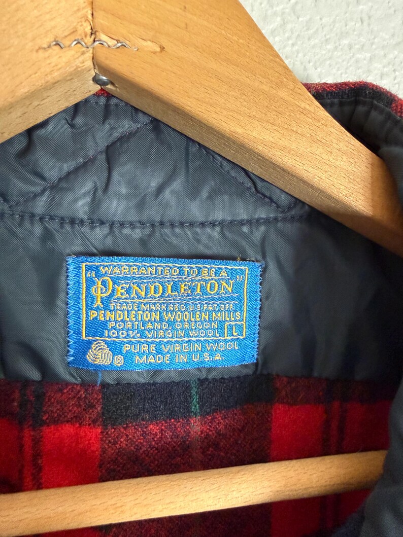 Puede incluir: Primer plano de una chaqueta de lana a cuadros rojos y negros. La chaqueta tiene un forro acolchado azul y una etiqueta azul que dice "WARRANTED TO BE A PENDLETON" y "PURE VIRGIN WOOL MADE IN U.S.A.". Cuelga de una percha de madera.