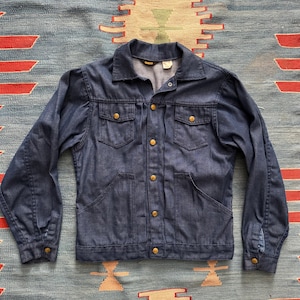 May include: A vintage denim jacket in a medium blue hue, featuring a classic collar, button-down front, and multiple pockets. The jacket has gold-colored buttons and a tailored fit, suggesting a retro style. The jacket is laid flat on a patterned rug.