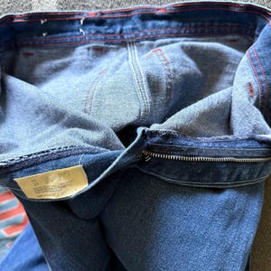 K&ouml;nnte beinhalten: Nahaufnahme einer blauen Jeans, die die Innenseite des Bunds und des Rei&szlig;verschlusses zeigt. Rote N&auml;hte akzentuieren die N&auml;hte. Ein Etikett mit Text ist sichtbar. Die Jeans ist umgeklappt und gibt den Innenstoff frei.