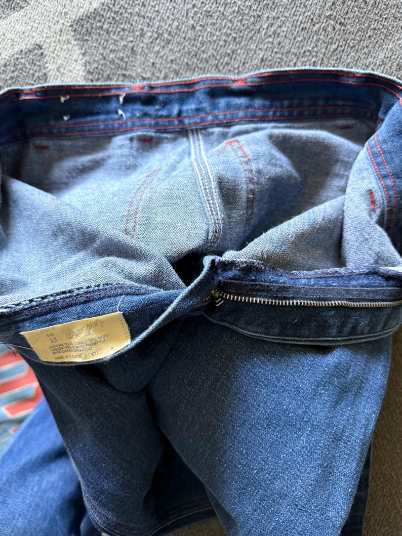 K&ouml;nnte beinhalten: Nahaufnahme einer blauen Jeans, die die Innenseite des Bunds und des Rei&szlig;verschlusses zeigt. Rote N&auml;hte akzentuieren die N&auml;hte. Ein Etikett mit Text ist sichtbar. Die Jeans ist umgeklappt und gibt den Innenstoff frei.