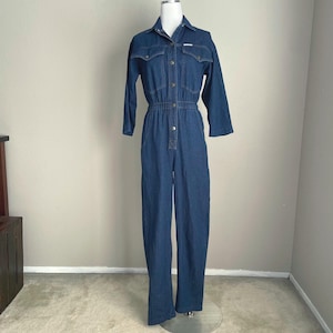 May include: A dark blue denim jumpsuit featuring a collared neckline, long sleeves, and a button-down front closure. The garment includes two chest pockets with flaps, an elasticized waistline, and straight-leg trousers in a classic indigo wash.