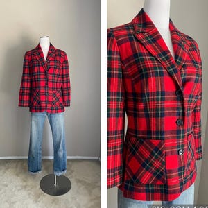 May include: A red, black, and green tartan plaid blazer with two black buttons and two front pockets. The blazer is displayed on a mannequin with blue jeans. The blazer's pattern is a classic tartan design.
